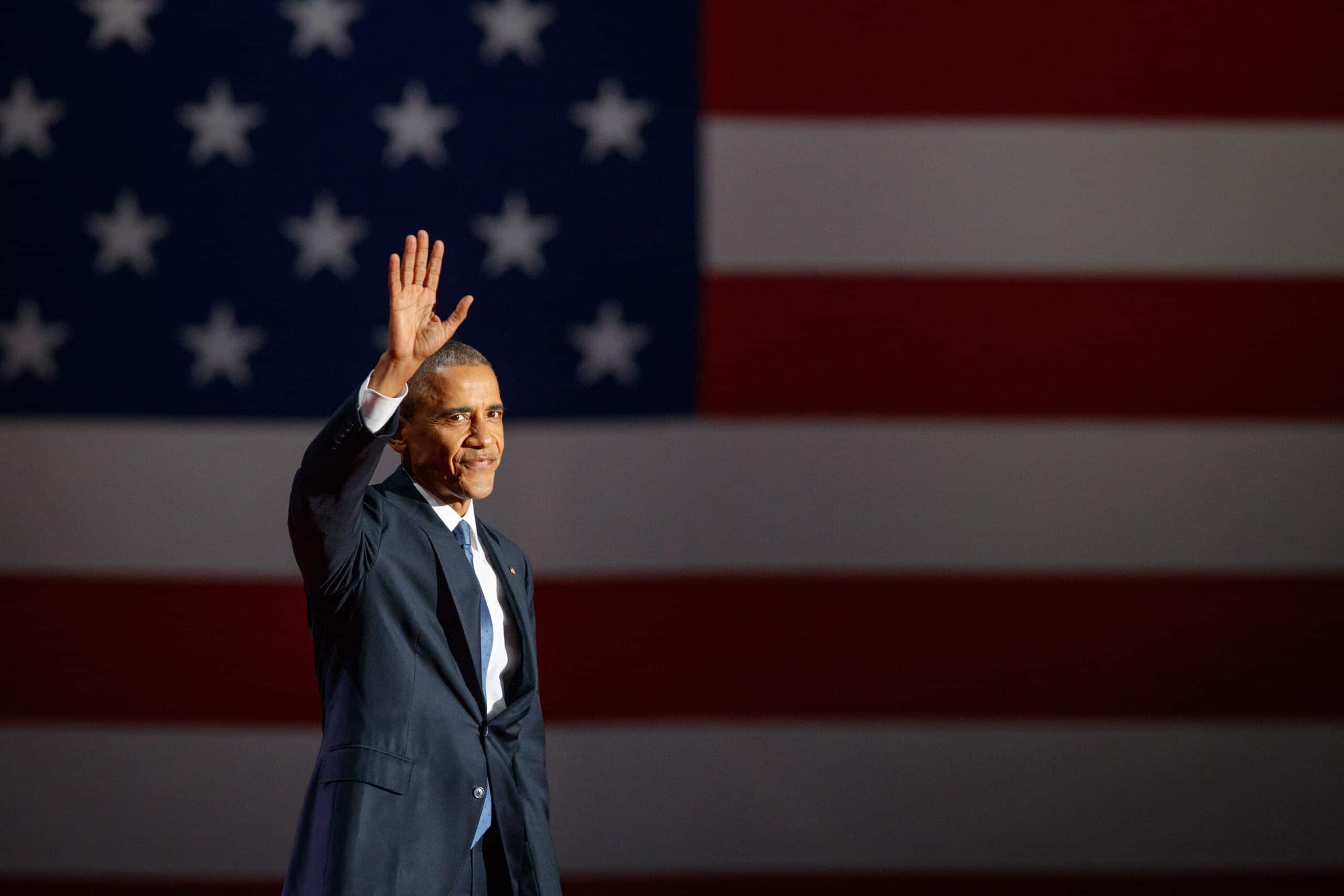 May contain one person wearing a dark suit and waving, with an American flag backdrop.