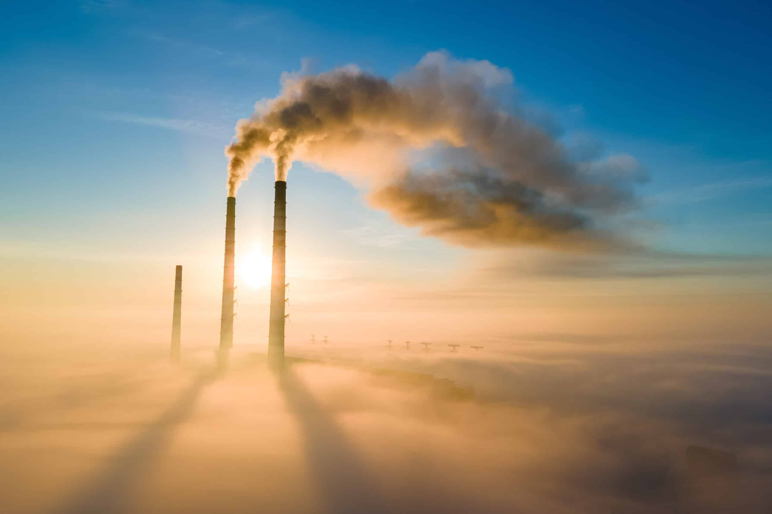 May contain smokestacks emitting smoke into foggy sky with sunrise.