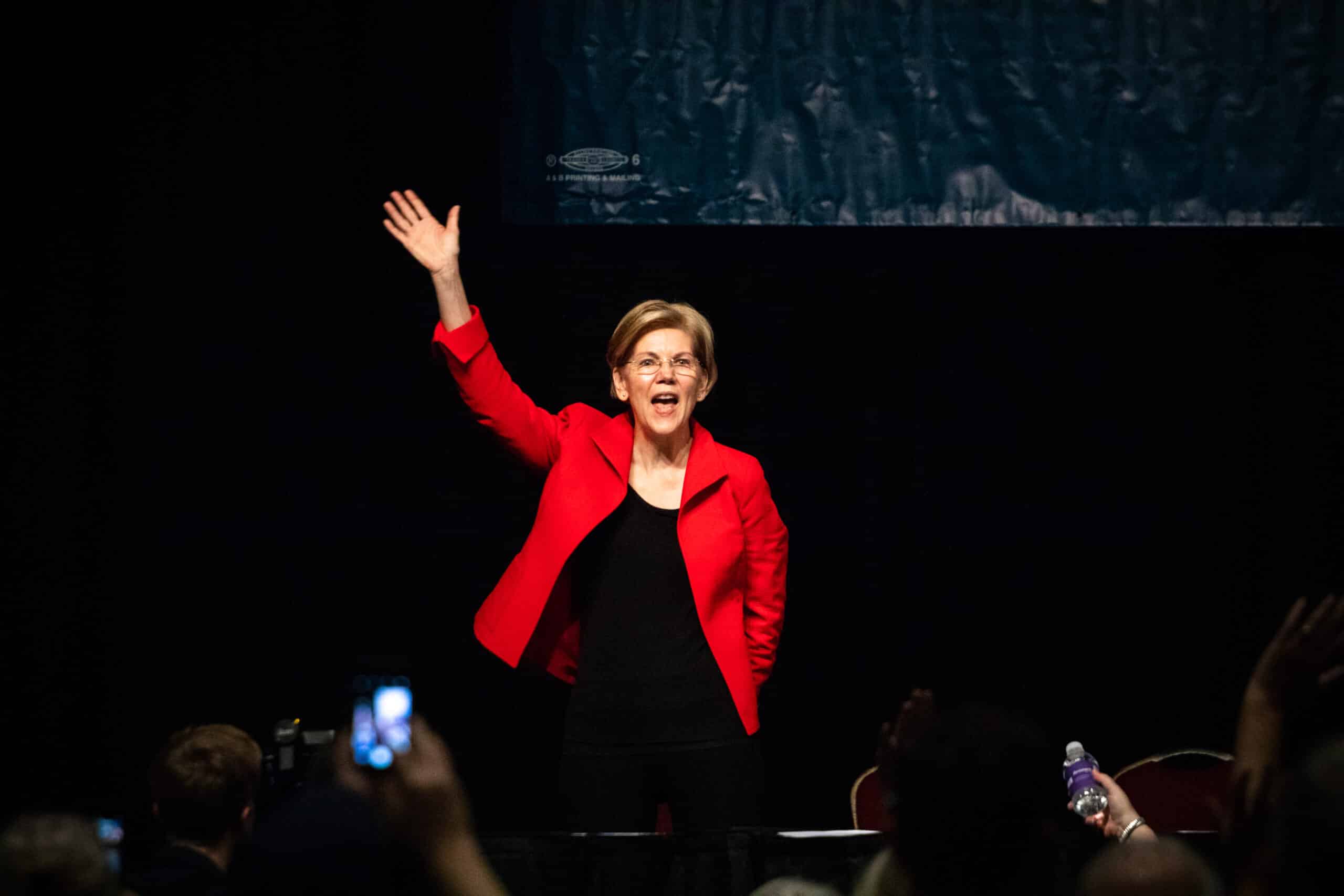 May contain one person in a red blazer raising a hand in front of a crowd.
