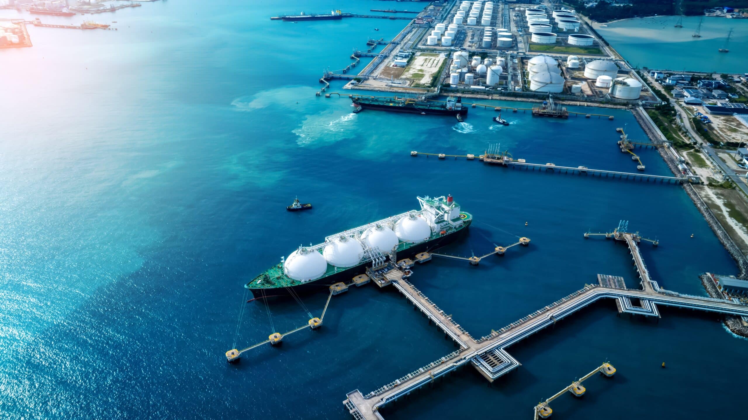 May contain an LNG tanker docked at an industrial port with storage tanks and blue water.