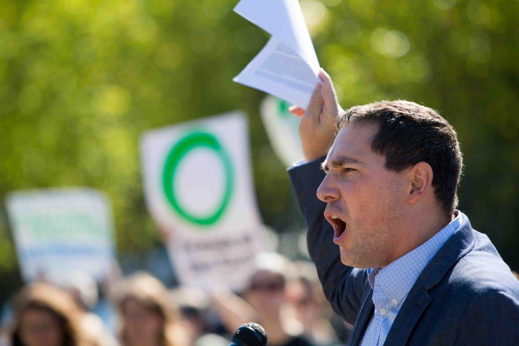 May contain a person speaking at a microphone holding papers.