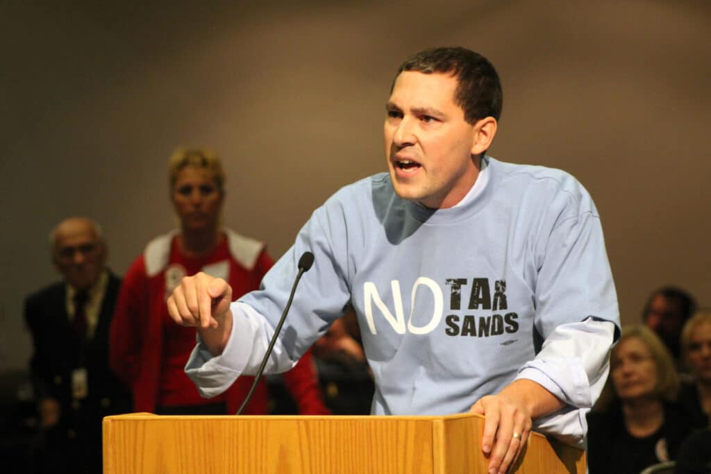 May contain a person speaking at a podium wearing a "No Tar Sands" shirt.