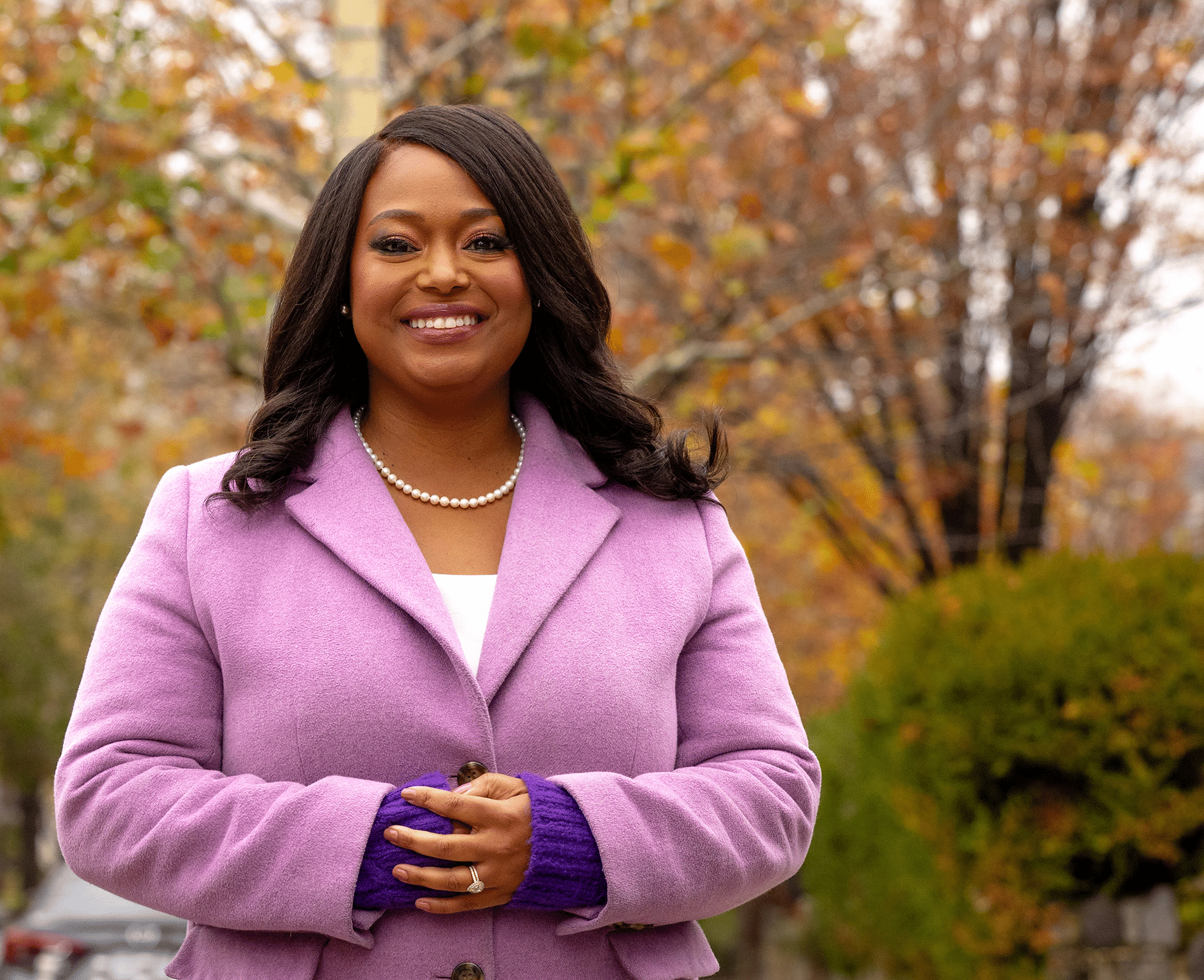 May contain one person in a lavender coat, smiling outdoors with fall foliage.
