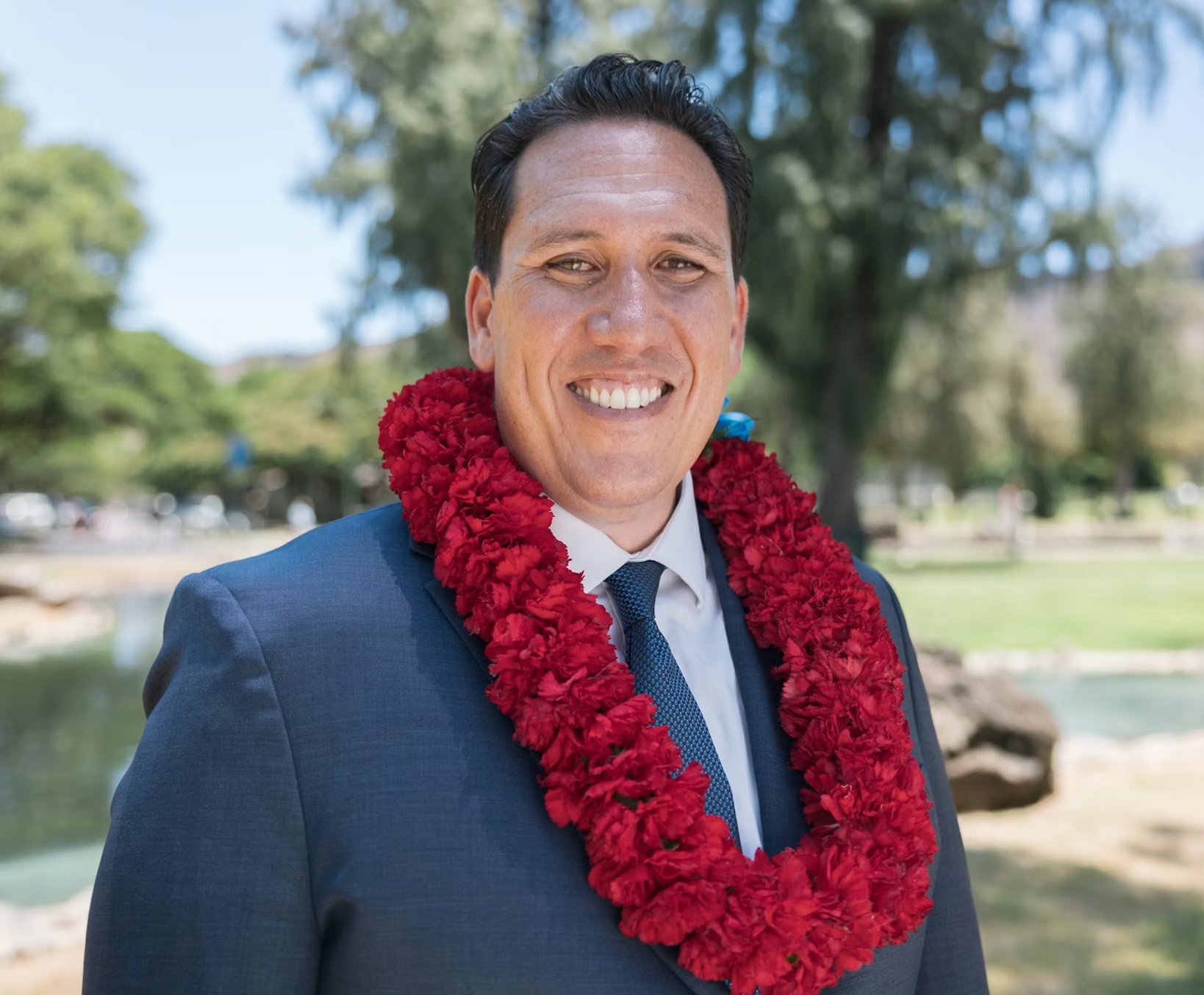 A man in a dark blue suit, white shirt, and blue tie stands outdoors, smiling with a red lei. Trees, grass, and a pond fill the sunny backdrop—perfect for candidate endorsements and community events.