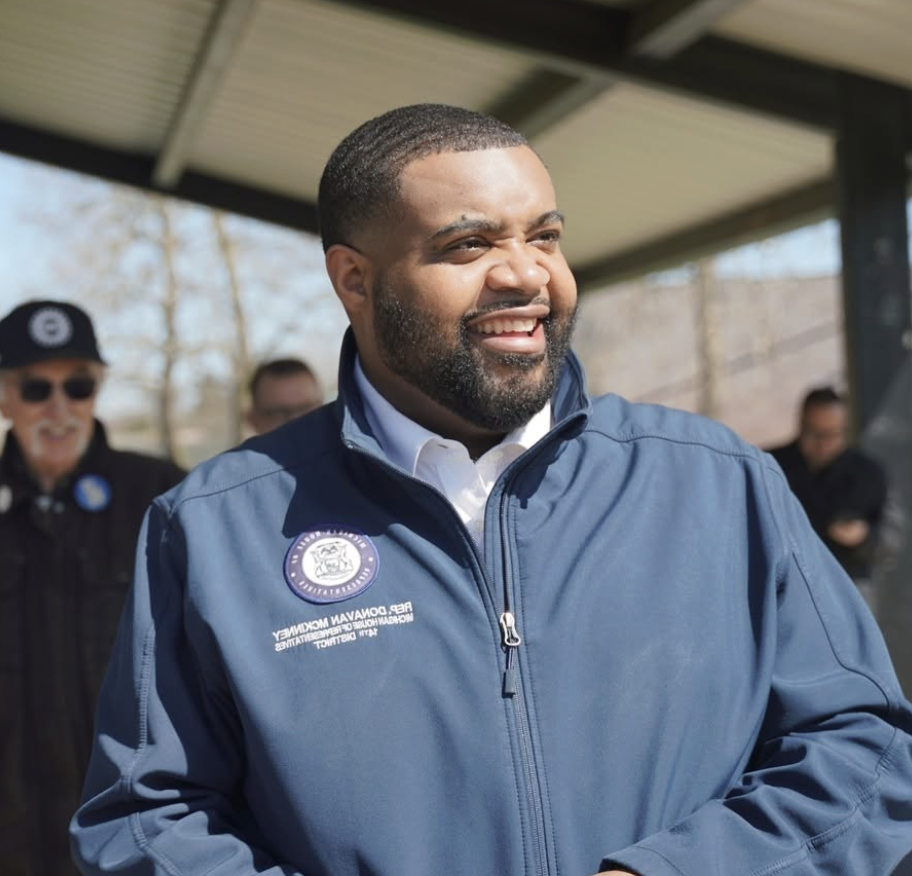 May contain a person in a blue jacket with a badge, smiling outdoors. Two people in background.